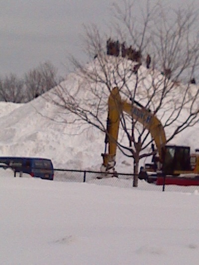 Earth moving equipment in front of a mountain of snow, topped by foolishly daring MIT students
